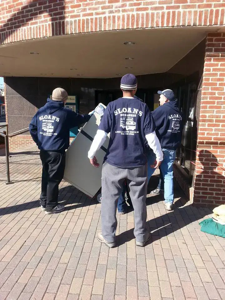 Uniformed Sloan's crew members carrying a padded item into a building